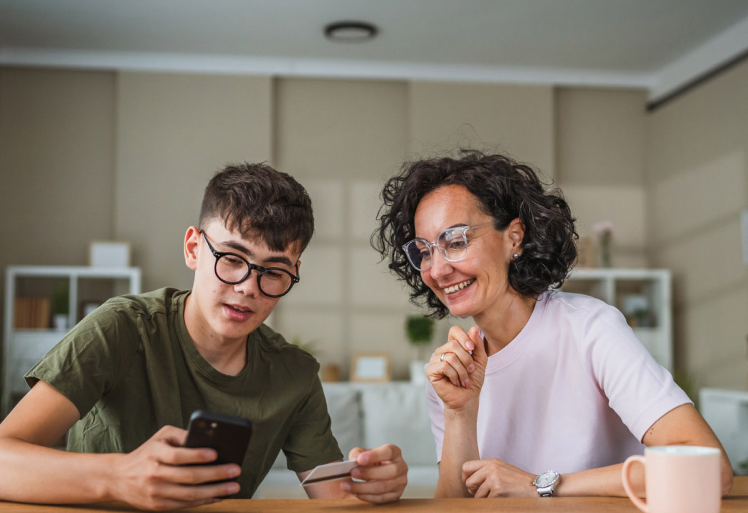 mom and teen are sitting at the table and looking at son's smartphone and plastinc card